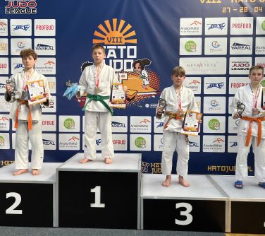 Sala. Na podium czterech chłopców w białych kimonach. W tle baner z logami