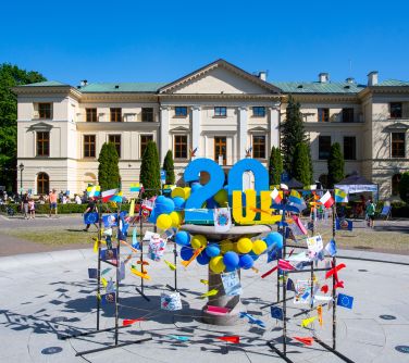 Na dworze. Na postumencie duża cyfra 20 niebieska. Naokoło na sznurkach kolorowe flagi, samoloty, laurki