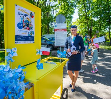 Na dworze. Przy żółtym wózku promocyjnym z tablicą stewardessa. W wózku niebieskie wiatraczki. Na tablicy plakat
