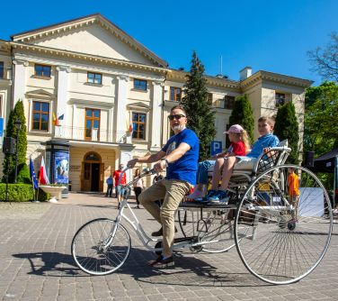 Dwór. Mężczyzna jedzie monocyklem. Z tyłu na siedzeniu chłopiec i dziewczynka. W tle pałac