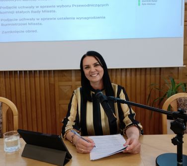 Sala. Uśmiechnięta kobieta siedzi przy długim stole przed mikrofonem i dokumentami. W tle ekran