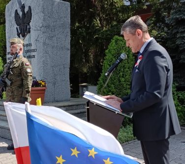 Na dworze. Przed mówinica przemawia mężczyzna w ciemnym garniturze. Z boku pomnik z kamienia. Przed nim żołnierz z bronią