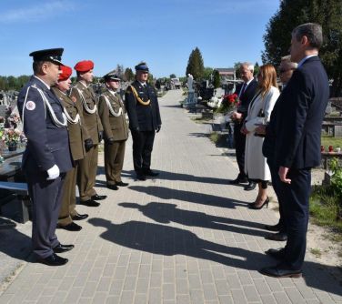 Cmentarz. Na chodniku na przeciwko siebie stoja osoby. Naokoło groby i drzewa