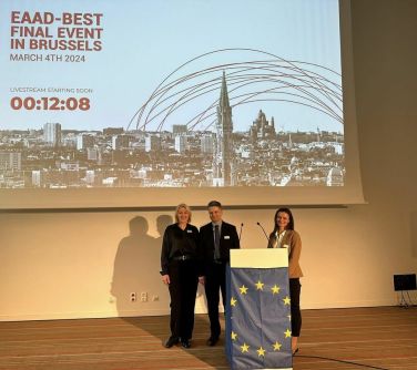 Aula. Trzy osoby stoja przed mównicą okalana flagą unii europejskiej. W tle wyświetlane zdjęcie miasta