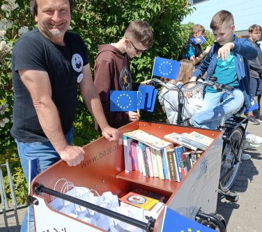 Na dworze. Uśmiechnięty mężczyzna stoi przy wózku wypełnionym książkami. Obok dzieci. W tle wysoki, zielony krzak