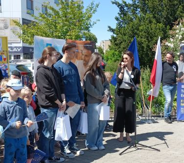 Na dworze. Kobieta przemawia do mikrofonu. Po lewej troje zwycięzców w konkursie oraz gromadka dzieci