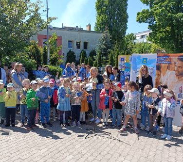 Na dworze. Gromadka dzieci z flagami Europy śpiewa. W tle roll-upy, drzewa  i osoby dorosłe