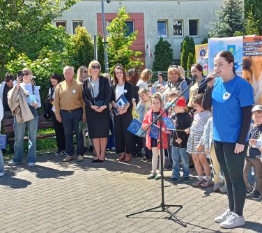 Na dworze. Nastolatka w niebieskiej koszulce śpiewa do mikrofonu. Naokoło osoby