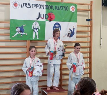 Sala gimnastyczna. Trzy dziewczynki w białych kimonach stoją na miejscach na podium z nagrodami i dyplomami w dłoniach. W...