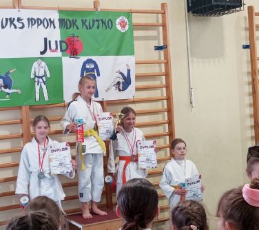 Sala gimnastyczna. Trzy dziewczynki w białych kimonach stoją na miejscach na podium, czwarta obok podium, z nagrodami i...