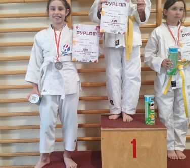 Hala. Trzy dziewczynki pozują stojąc na podium z nagrodami w dłoniach. W tle baner na drabinkach