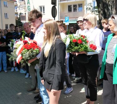 Na dworze. Zgromadzone osoby. Szpaler po dwie osoby trzymają wiązanki z biało-czerwonych kwiatów