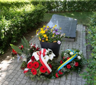Na dworze. Tablica upamiętniająca. Za nią trawnik i krzewy. Przed nia donica z żółto-fioletowymi bratkami i trzy wiązanki z...