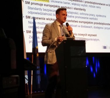 Pomieszczenie, scena. Mężczyzna przemawia przed mównicą. W tle na ścianie na ekranie wyświetlana treść