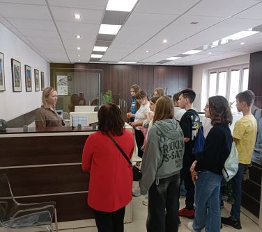 Sala. Grupa nastolatków słucha stojącej za szybą kobiety