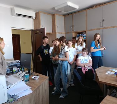 Pokój. Grupa nastolatków słucha opowieści kobiety stojącej za biurkiem