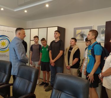 Biuro. Grupa nastolatków słucha stojącego przed nimi mężczyzny w błękitnej marynarce. Obok trzy czarne fotele. W tle roll-up