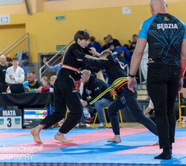 Hala sportowa. Dwóch chłopców walczy na kolorowych matach. Obok sędzia