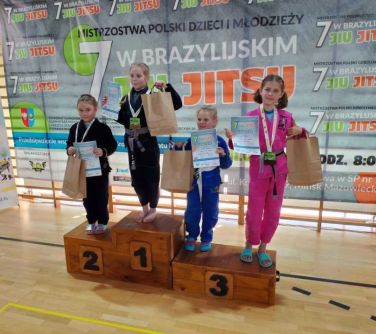 Hala sportowa. Cztery dziewczynki w kolorowych kimonach z nagrodami w rękach stoją na podium. Dwie na miejscu trzecim. W...