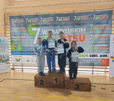 Hala sportowa. Czwórka dzieci z nagrodami w rękach stoją na podium. Dwie na miejscu pierwszym. W tle baner na drabinkach