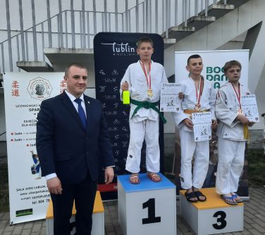 Pomieszczenie. Trzech zawodników w białych kimonach na podium. Po lewej stoi mężczyzna w ciemnym garniturze