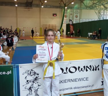 Hala sportowa. Dziewczynka z pucharem i dyplomem stoi przed barierką. Za nią kolorowe, piankowe maty na których walczą...