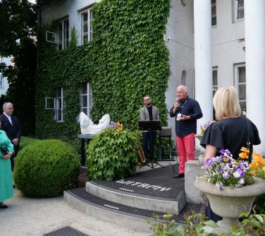Na zewnątrz. na schodach wejściowych do budynku z filarami przemawia do mikrofonu mężczyzna w ciemnej koszuli. Po lewej dom...