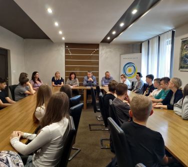 Sala konferencyjna. Przy stołach ustawionych w literę U na krzesłach siedzą osoby