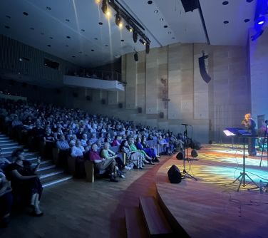 Sala koncertowa. Po prawej na scenie śpiewa kobieta. Po lewej widownia