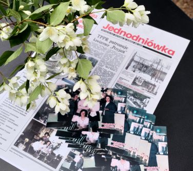 Na czarnym obrusie leży stara gazeta jednodniówka, na niej stare pocztówki. Po lewej wazon z jaśminem