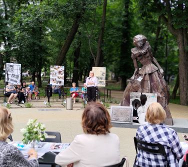 Dwór. Przed pomnikiem kobiety z mosiądzu osoby siedzą. W tle kobieta przemawia i osoby siedzą na ławkach