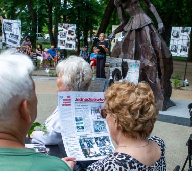 Dwór. Kobieta siedząc czyta gazetę jednodniówkę. Obok osoby. W tle pomnik kobiety z mosiądzu i pozsotałe osoby