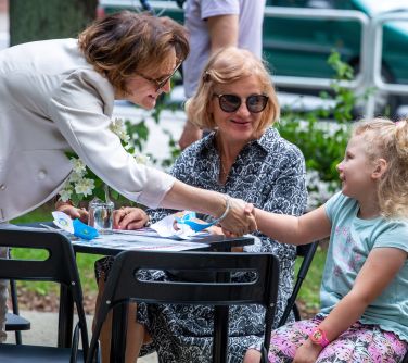 Dwór. Kobieta pochyla się nad stołem i podaje rękę siedzącej na przeciw dziewczynce. Obok druga kobieta przygląda się siedząc