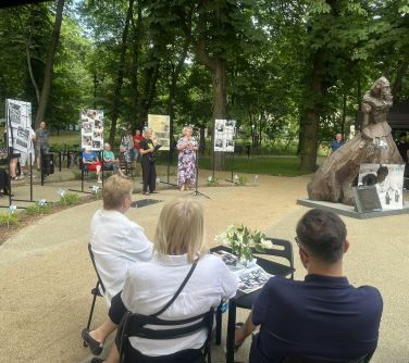 Dwór. Trzy osoby siedzą przy stoliku. Słuchają przemawiającej na przodzie kobiety. Po prawej pomnik kobiety z mosiądzu