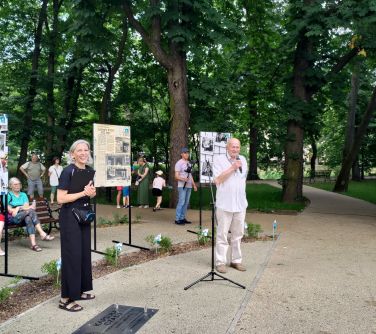 Dwór. Mężczyzna w jasnym ubrania przemawia do mikrofonu. Po lewej uśmiechnięta kobieta. W tle wystawa i osoby