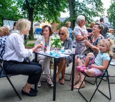 Dwór. Przy stole siedzą osoby, rozmawiają i jedzą. Naokoło stoją osoby.
