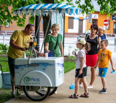 Dwór. Saturator do wody. Mężczyzna nalewa wodę. Osoby stoją w kolejce