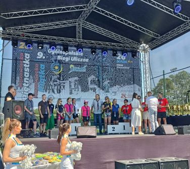 Scena na zewnątrz. Na scenie osoby z krawatami na szyjach. Po prawej na stole złote puchary.