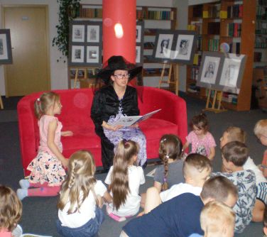 Sala biblioteczna. Na czerwonej sofie siedzi kobieta w czarnym kapeluszu. Czyta zgromadzonym przed nią dzieciom