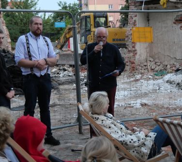 Na dworze, na ruinach domu dwóch mężćzyzn, jeden przemawia. Na przodzie siedzą na leżakach