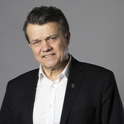 Zdjęcie portretowe. Mężczyzna uśmiecha się. Ma na sobie białą koszulę i czarną marynarkę. Za nim szare tło.