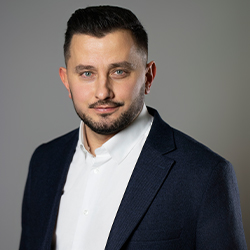 Zdjęcie portretowe. Mężczyzna delikatnie się uśmiecha. Ma na sobie białą koszulę i granatową marynarkę. Za nim szare tło.