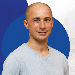Zdjęcie portretowe. Mężczyzna w szarym swetrze uśmiecha się. Biało-granatowe tło.
