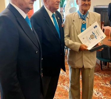 Trzech mężczyzn w garniturach. Pozują do zdjęcia, jeden z nich trzyma w rękach dyplom. Ma na szyi medal.