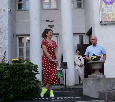 Dzień. Na schodach pałacu między filarami stoi mężczyzna i dwie kobiety. Na froncie kobieta w czerwonej sukience w białe...