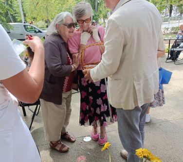 Skwer, w ciągu dnia. Mężczyzna w jasnej czekoladzie częstuje dwie kobiety giftem z koszyka