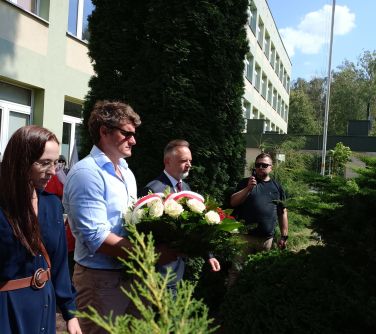 Dwór. Dwóch mężczyzn i kobieta skłądają biało-czerwoną wiązankę przed pomnikiem. Naokoło krzewy. W tle budynek szkoły