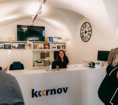 Pomieszczenie. Za białą ladą siedzi kobieta. W tle na półkach ksiązki, po prawej na białej ścianie czarny zegar