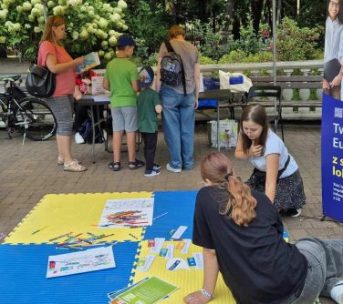 Dzień. Dwie nastolatki siedzą na żółto-niebieskich piankach na podłodze. W tle stoją osoby