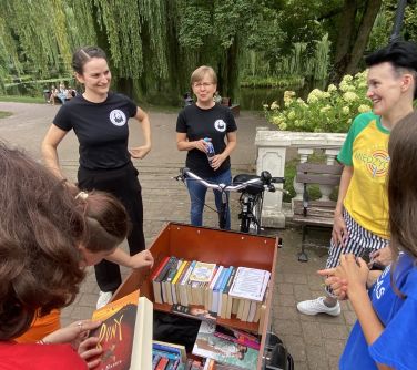 Dzień, park. Stoi kilka uśmiechniętych osób. Na przodzie półka z książkami